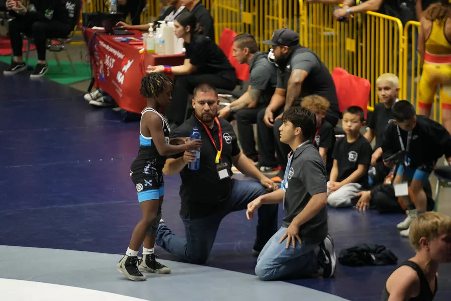 A coach kneeling matside to talk with a young Deep Waters wrestler between periods